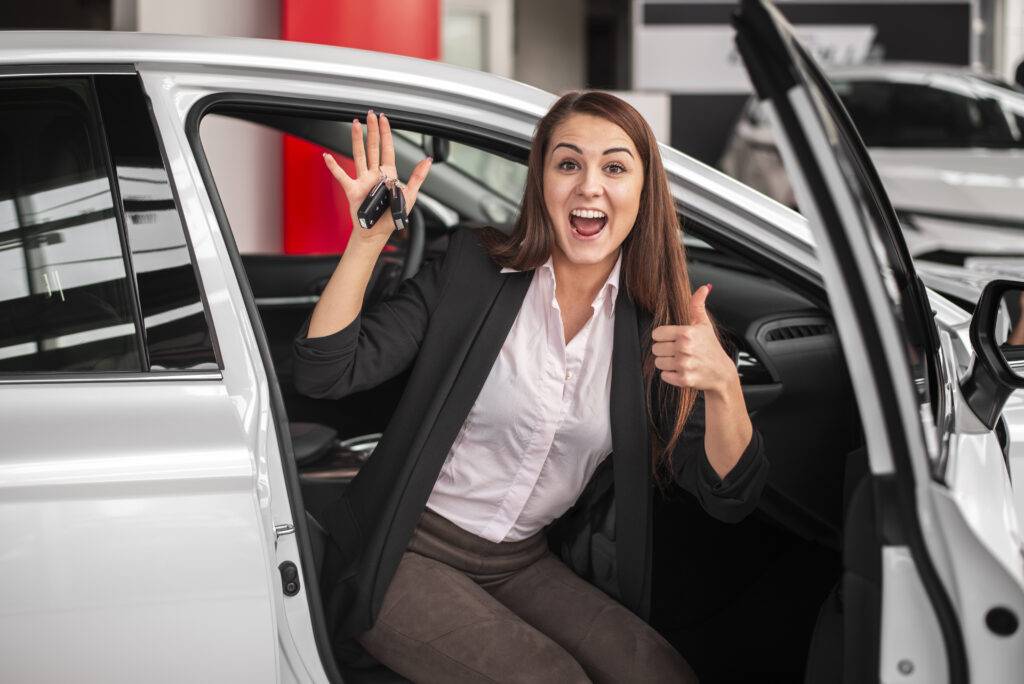 Excited woman holding car keys and giving a thumbs up by a new car.